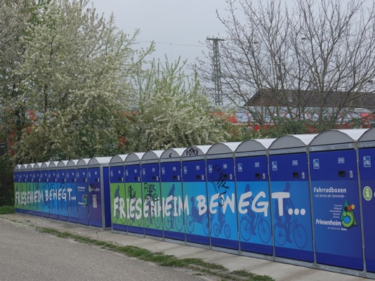 Foto: mehrere blaue Radboxen (abschließbare Fahrradgaragen) in einer Reihe, im Hintergrund Hecken und dahinter das Dach eines Zugwaggons