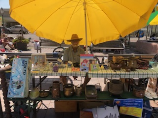 Mann mit Strohhut und Bart sitzt unter einem schräggestellten orangegelben Sonnenschirm hinter einem großen Tisch und eine Bank mit Flohmarkt-Dingen zum Verkaufen und Verschenken.