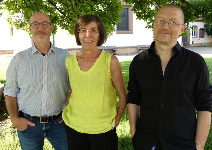 Drei Menschen unter einem Baum: von links ein Mann (Stefan) in hellblauem Hemd und blauer Jeans, mit Brille, Stoppelbart einer Glatze, dann eine Frau (Simone) in gelbem, ärmellosem Shirt, schwarzer Hose, rahmenloser Brille, glatten, braunen Haaren, dann ein Mann (Michael) in schwarzem Hemd, mit Nickelbrille, Stoppelbart und sehr kurzen Stoppelhaaren. Alle drei lächeln und haben die Hände in den Hosentaschen.